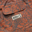 Close-up of rust-orange and dark gray patterned boardshorts with a pocket. The pocket features a sewn white Howler Brothers label with a palm tree graphic, two small orange eyelets at the bottom, and an elastic waistband for comfort.