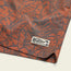 Close-up of a rust-orange fabric with a dark leaf and plant pattern, featuring a stitched Howler Brothers brand tag on the hem—perfect for boardshorts with an elastic waistband.