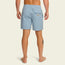 A barefoot person stands facing away, wearing light blue patterned boardshorts with an elastic waistband, a back pocket, and a small rectangular patch label. The person has a tattoo on their left calf. The background is plain and light-colored.