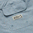 Close-up of a light blue fabric with small white, orange, and gray dots, featuring a flap pocket and a Howler Brothers label with a palm tree illustration—crafted in boardshorts style with an elastic waistband for comfort.
