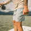 A person stands on a boat, holding a fishing rod and line, wearing light blue boardshorts with an elastic waistband and Howler patch, plus a patterned long-sleeve shirt. Water and mountains stretch across the background.