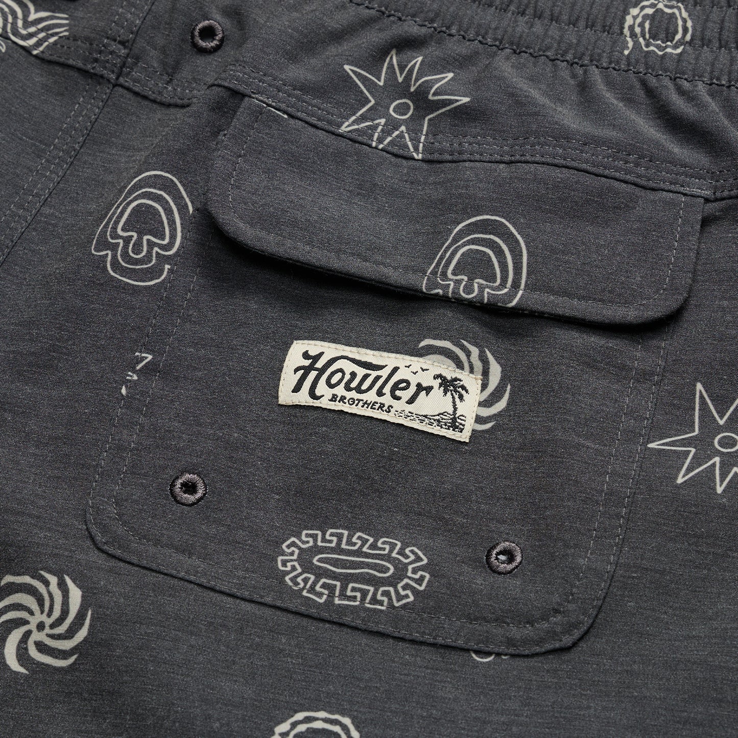 Close-up of a dark gray boardshorts pocket with abstract white patterns and a stitched Howler Brothers label under a palm tree graphic. The pocket, part of 7” inseam shorts, features two button holes at the bottom.