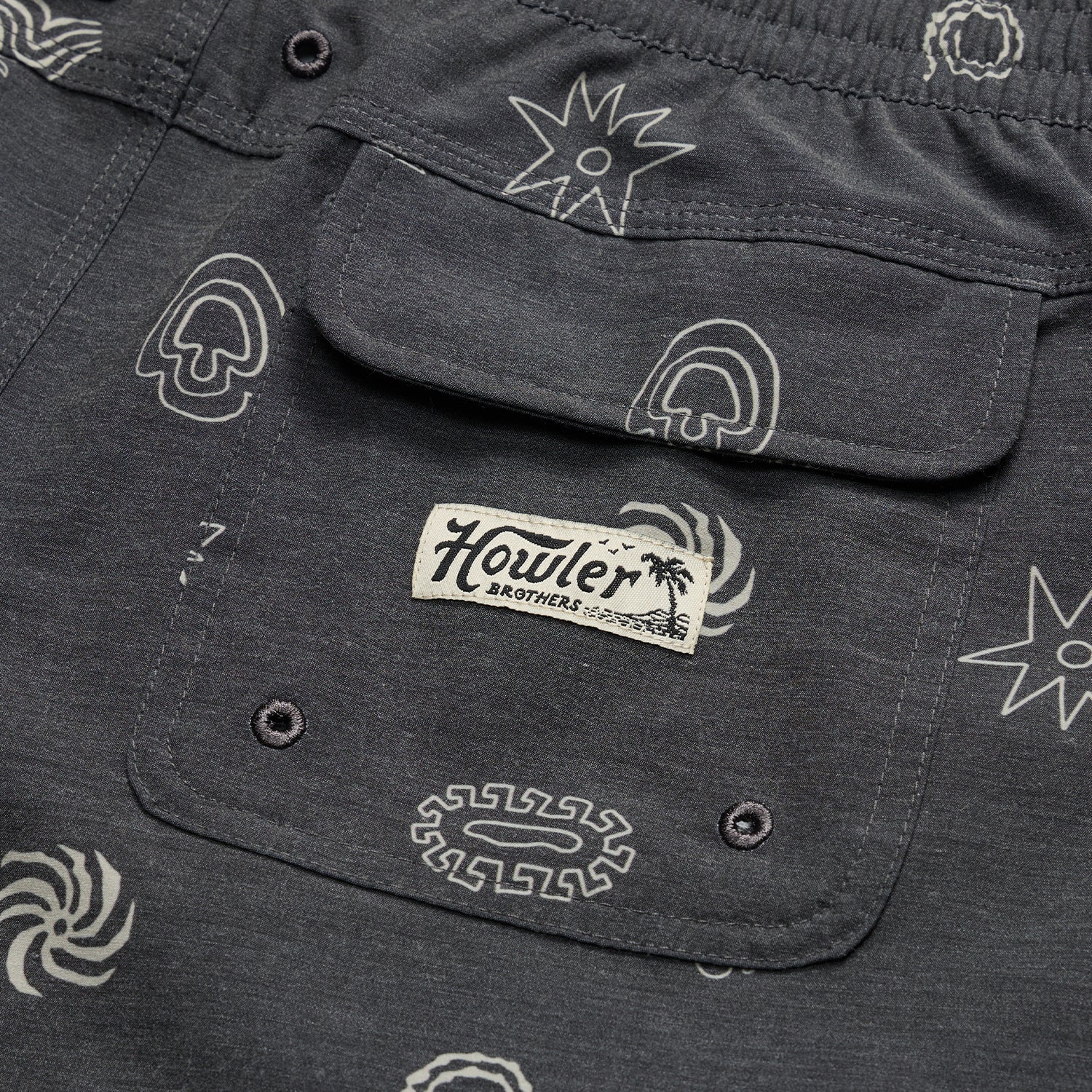Close-up of a dark gray boardshorts pocket with abstract white patterns and a stitched Howler Brothers label under a palm tree graphic. The pocket, part of 7” inseam shorts, features two button holes at the bottom.