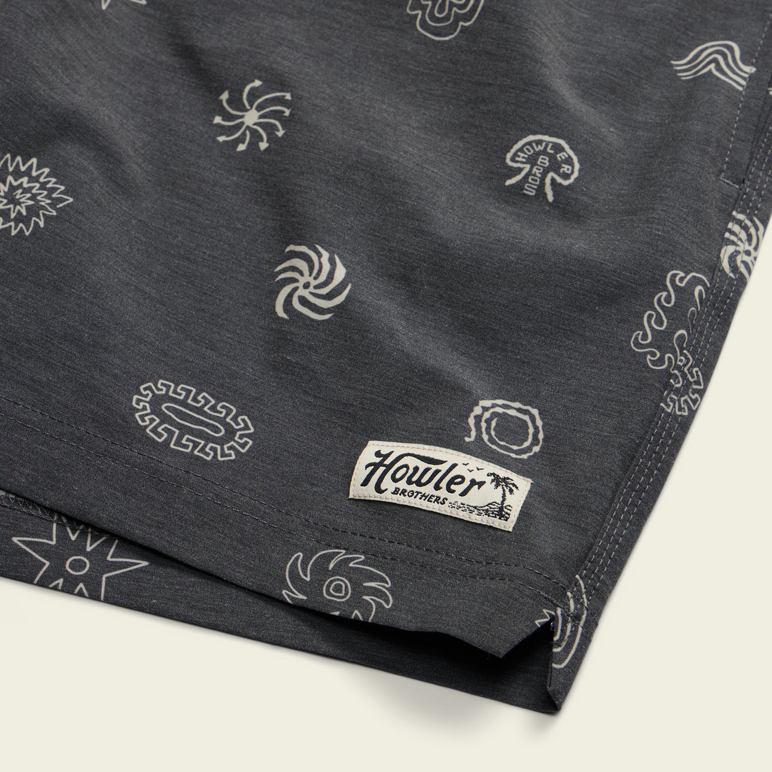 Dark boardshorts with white abstract and sun-like designs, featuring a 7” inseam and an elastic waistband. Finished with a small rectangular Howler Brothers label with a palm tree near the hem.