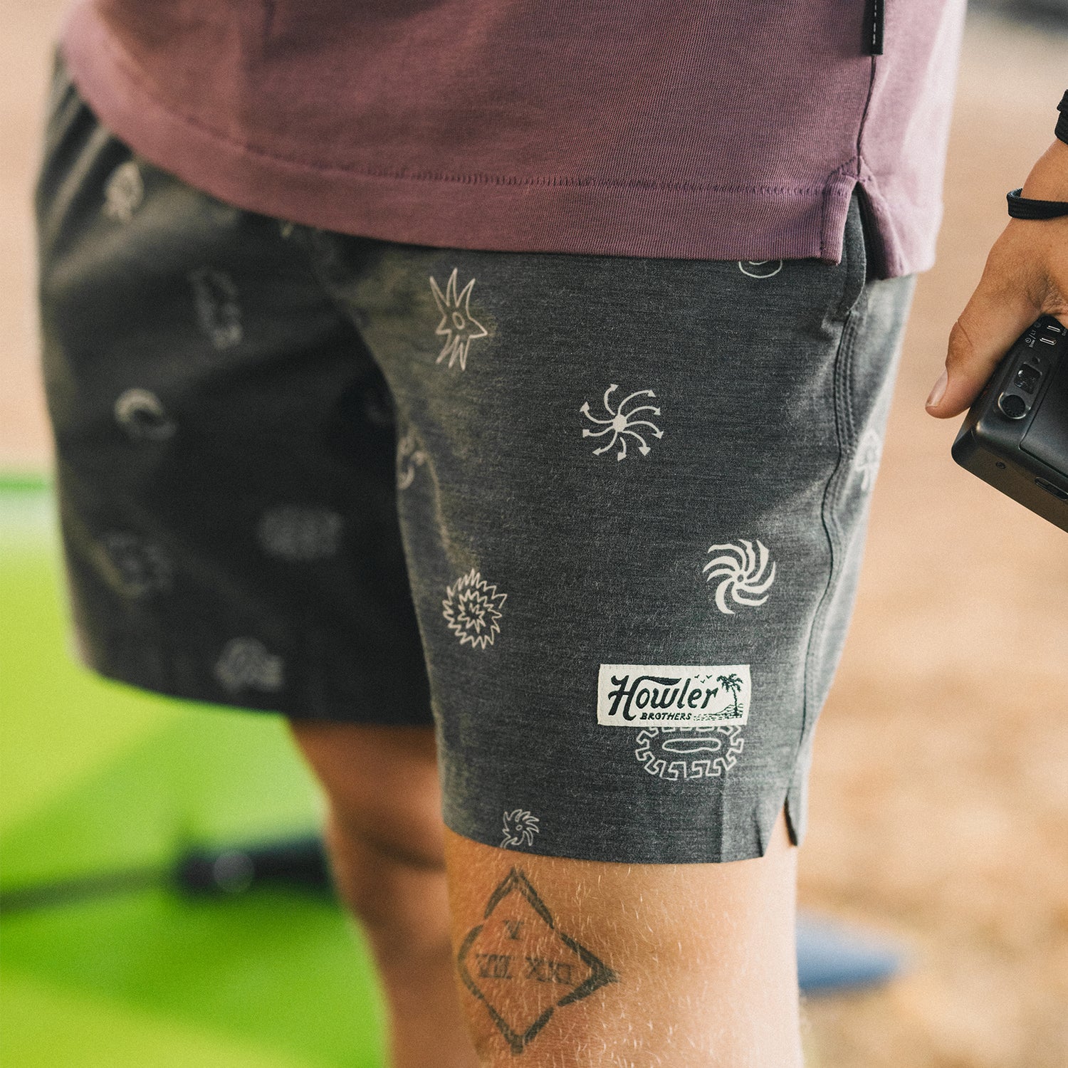 A person stands outdoors wearing boardshorts with a 7” inseam, featuring white sun and wave designs on dark fabric. Part of a purple shirt is visible, and they are holding a black object in their right hand.