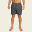 A man wearing navy blue boardshorts with white printed patterns and a beige drawstring stands barefoot against a plain light background. These boardshorts feature an elastic waistband and a comfortable 7” inseam.