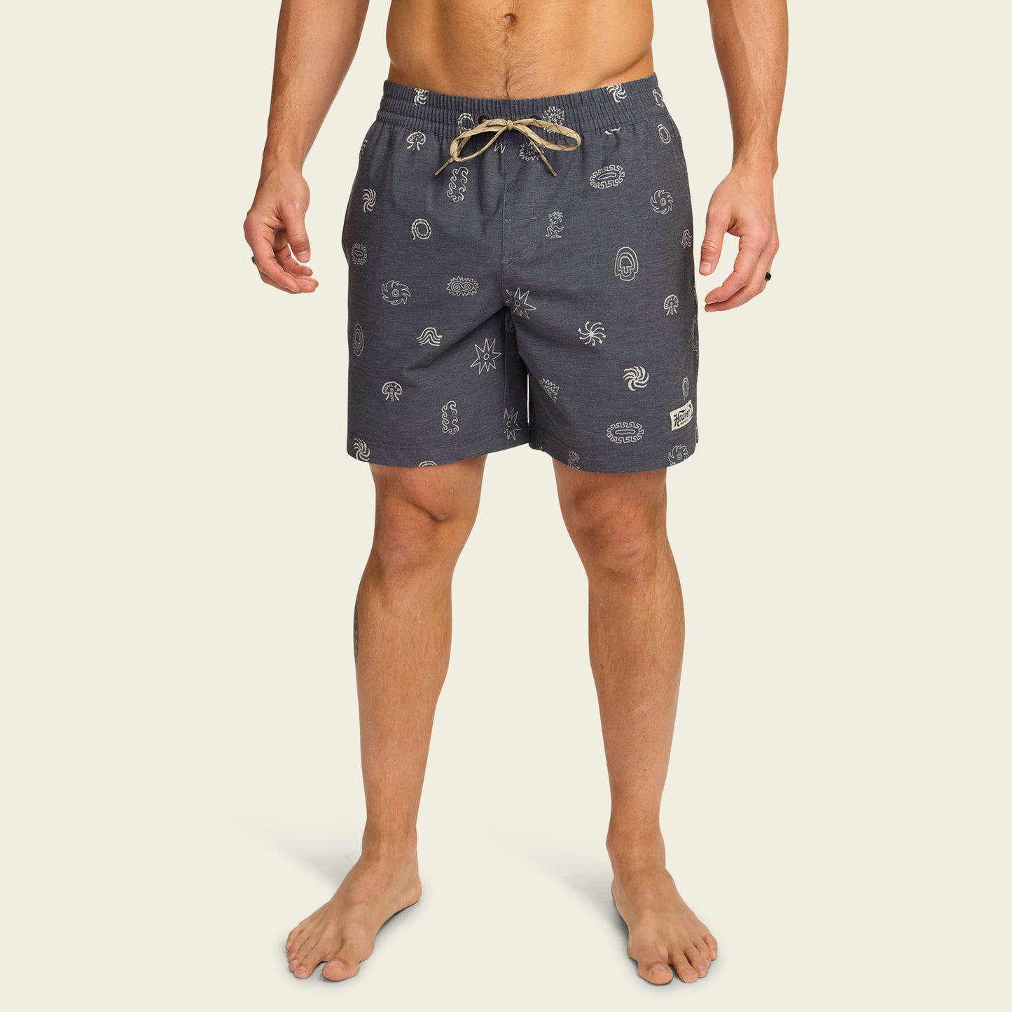 A man wearing navy blue boardshorts with white printed patterns and a beige drawstring stands barefoot against a plain light background. These boardshorts feature an elastic waistband and a comfortable 7” inseam.