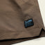 Close-up of a textured brown fabric, with a small black and blue Howler Brothers logo patch sewn near the edge and a side slit at the hem—perfect details for tech seersucker shorts.