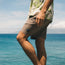 A person wearing a floral shirt and tech seersucker shorts stands by the ocean, with their hand resting on their thigh and blue water and a distant coastline visible in the background.