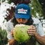 A man with long hair and a beard drinks from a green coconut while wearing a blue breathable tech hat with a fish graphic and the word “Howler.” He is outdoors, surrounded by lush tropical plants, ready for outdoor adventures.