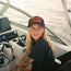 A woman with long blonde hair and tattoos, wearing a black T-shirt and a vintage-inspired high crown hat with a foam front, sits at the helm of a boat, smiling and looking to the side. The background shows calm water.