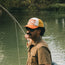 A man wearing sunglasses and a vintage-inspired hat holds a fishing rod while standing near the edge of a calm river, with trees and greenery in the background.