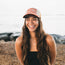 A woman with long brown hair wearing a vintage-inspired Howler Brothers cap and a black strapless top smiles outdoors near a rocky beach, with the ocean blurred in the background.