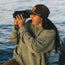 A person wearing a brown Unstructured Snapback with an adjustable snapback closure and green sweatshirt smiles while filming with a vintage video camera near a body of water.
