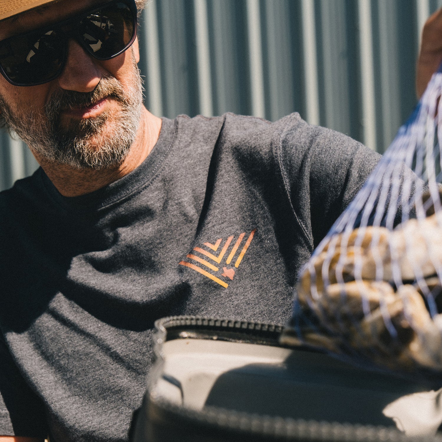 A man wearing sunglasses, a hat, and a JT Van Zandt Collection Howler Select T-Shirt with a geometric design holds a mesh bag filled with what appears to be potatoes outdoors near a corrugated metal wall.
