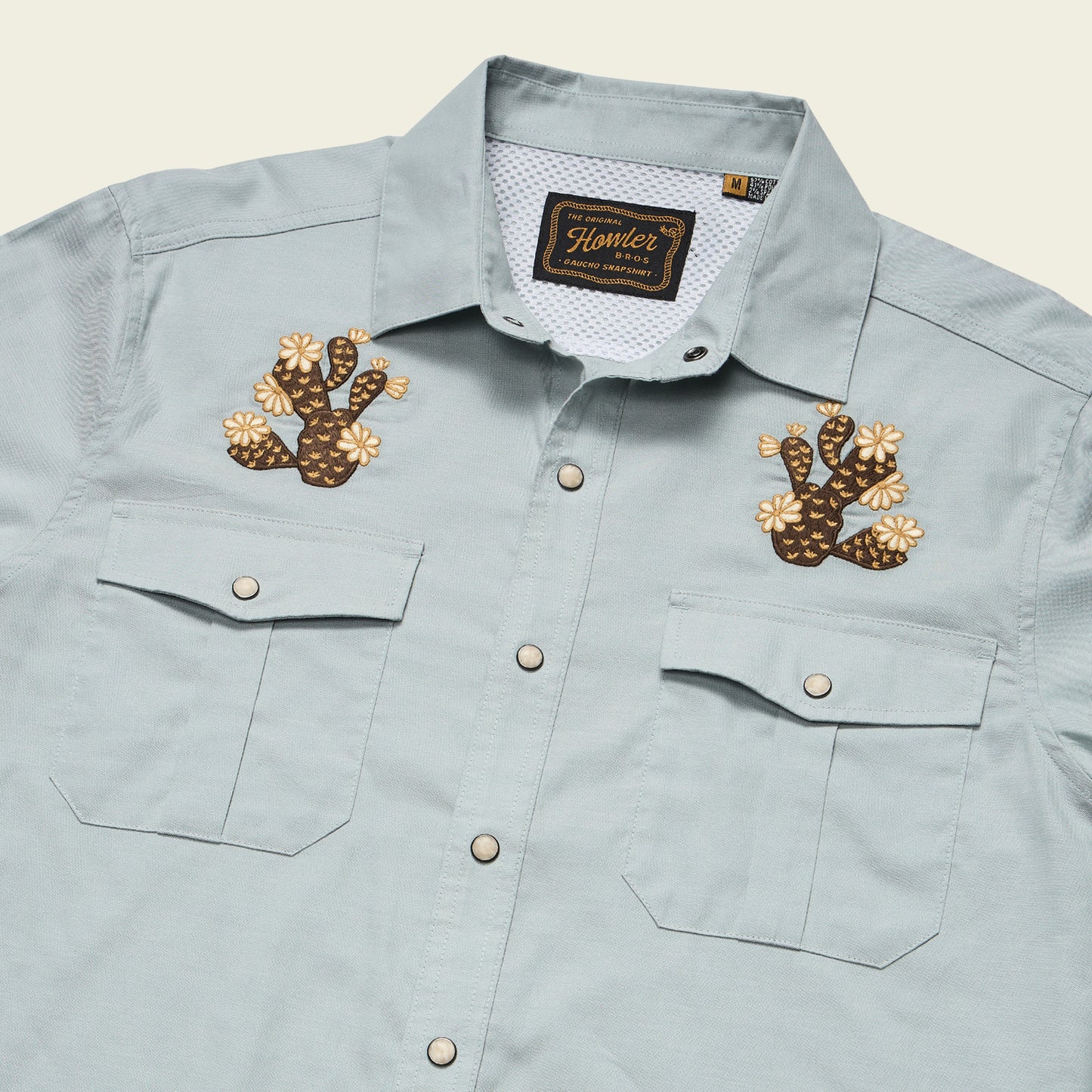 Light blue Gaucho Snapshirt with two front pockets and decorative cactus and flower embroidered artwork on each side of the chest. The shirt label reads Howler and the size is marked as medium, inspired by vintage western shirts.
