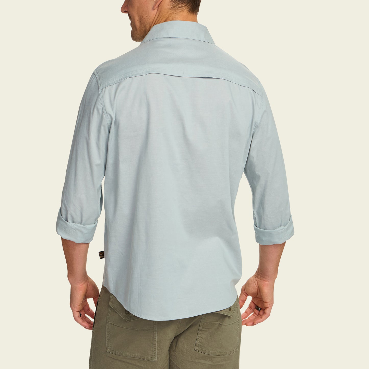A man wearing a light blue Gaucho Snapshirt with rolled-up sleeves and olive green pants is shown from the back against a plain light background.