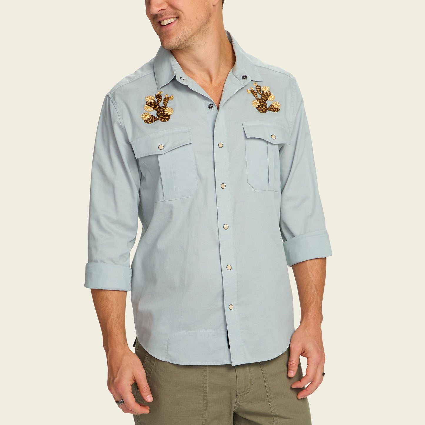 A man wearing a light blue Gaucho Snapshirt with rolled-up sleeves and decorative bear paw patches reminiscent of vintage western shirts, paired with olive green pants.