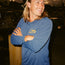 A smiling person with long blond hair, wearing a blue UPF 50+ hooded tech shirt with Howler written on it, stands outdoors with their arms crossed, near boats at a marina during sunset.