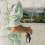A person wearing a light Matagorda Shirt is holding a fishing rod by a body of water, with greenery and mountains visible in the blurred background. This travel shirt is perfect for outdoor adventures.