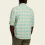 A person is shown from the back wearing a long-sleeve, light blue and cream plaid Matagorda Shirt with olive green pants. The shirt sleeves are rolled up, highlighting this quick drying travel shirt style as they keep their hands in their pockets.