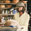 A bearded man in a cap, sunglasses, and a Matagorda Shirt uses a lathe to shape a large wooden bowl, with wood shavings flying in the quick-drying travel shirt, surrounded by tools and shelves in his busy workshop.