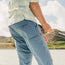 A person wearing a light plaid shirt and blue tech pants stands by the water, holding a fishing rod, with a blurred natural landscape and bright sky in the background.