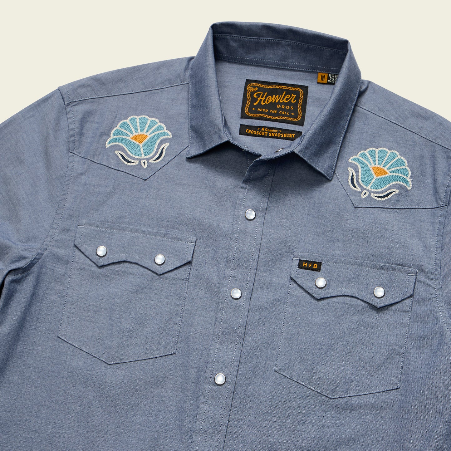 The Crosscut Deluxe Shortsleeve Shirt - Fresh Blooms: Indigo features snap buttons, two chest pockets, and floral embroidery on the shoulders. The label reads Howler and Honest Apparel.