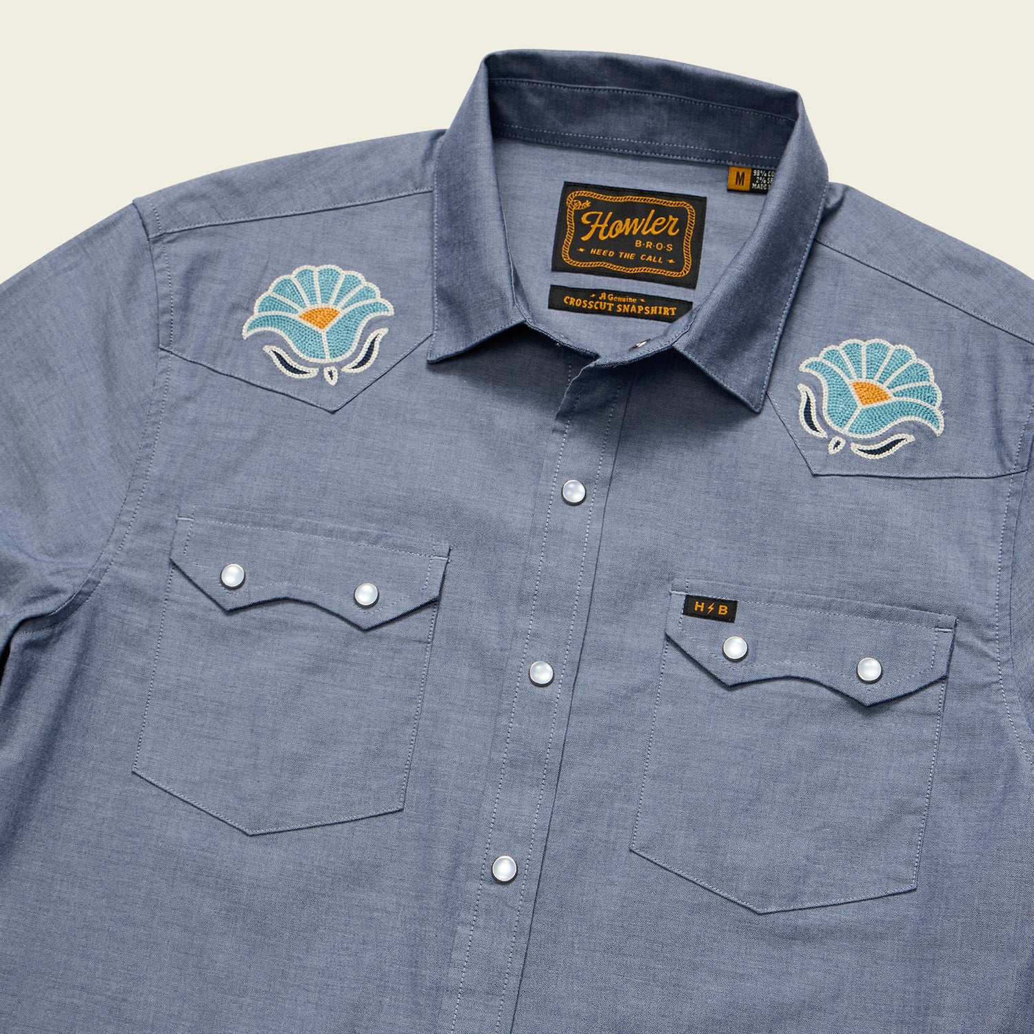 The Crosscut Deluxe Shortsleeve Shirt - Fresh Blooms: Indigo features snap buttons, two chest pockets, and floral embroidery on the shoulders. The label reads Howler and Honest Apparel.