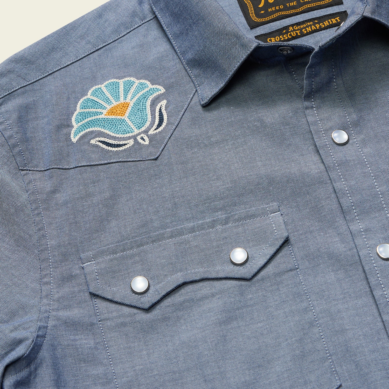 A close-up of a blue denim western-style Crosscut Deluxe Shortsleeve summer shirt with pearl snap buttons, two chest pockets, and an embroidered blue and white flower motif embroidery with an orange center on the left shoulder yoke.