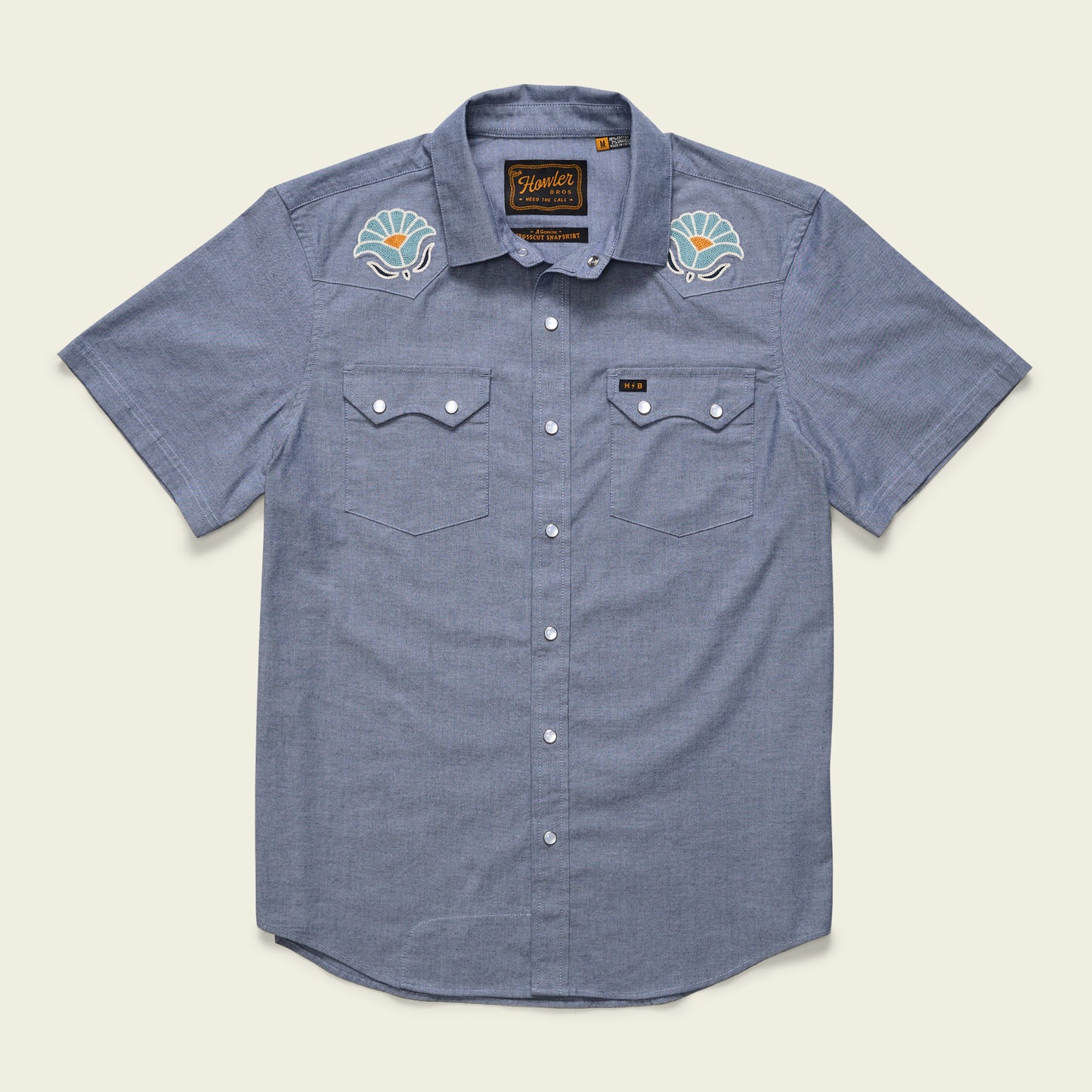 The Crosscut Deluxe Shortsleeve Shirt - Fresh Blooms: Indigo is a light blue summer shirt with two chest pockets, white buttons, and colorful seashell motif embroidery on the shoulders.