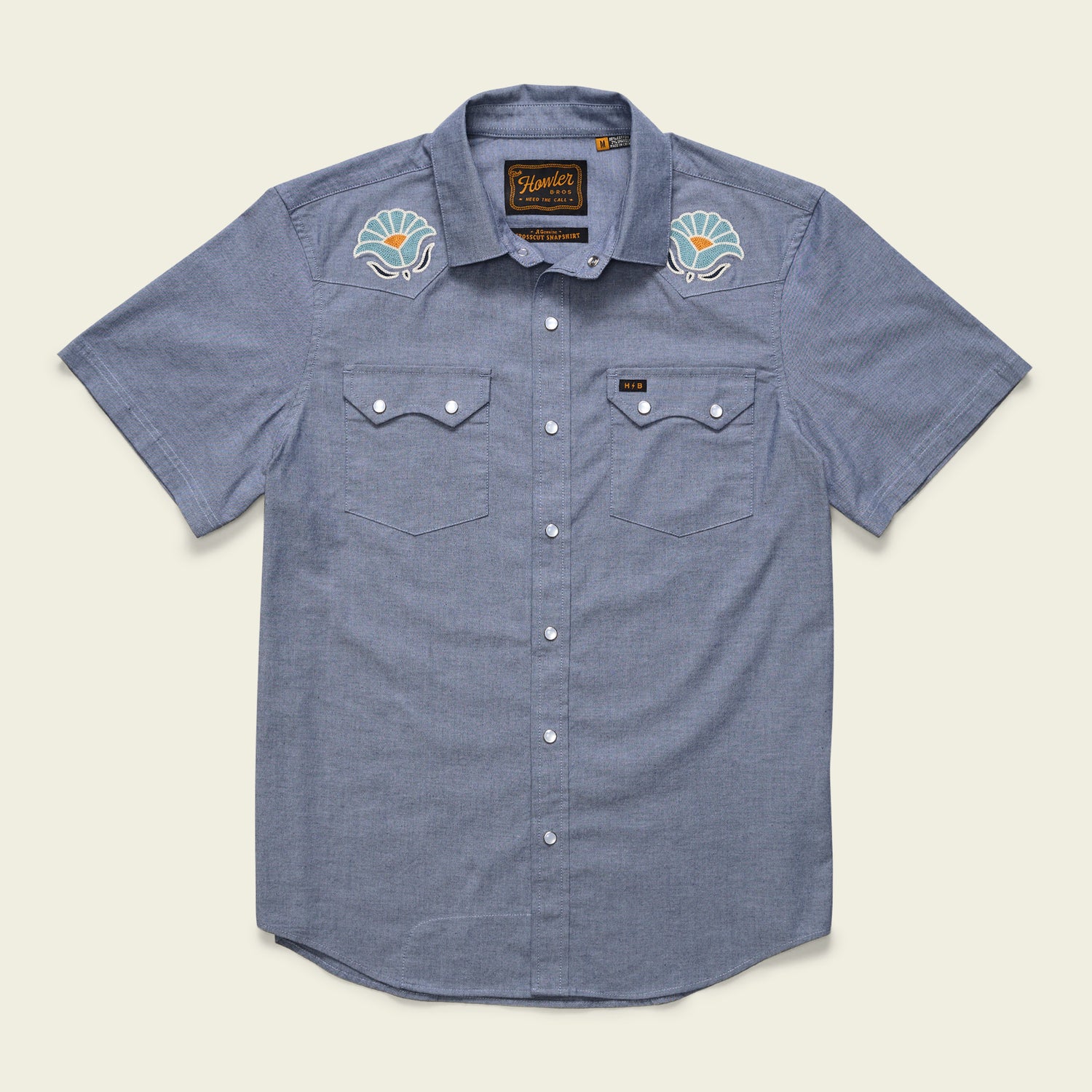 The Crosscut Deluxe Shortsleeve Shirt - Fresh Blooms: Indigo is a light blue summer shirt with two chest pockets, white buttons, and colorful seashell motif embroidery on the shoulders.