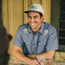 A man in a Crosscut Deluxe Shortsleeve Shirt - Fresh Blooms : Indigo and a trucker hat smiles while sitting at an outdoor wooden table with a drink in front of him.