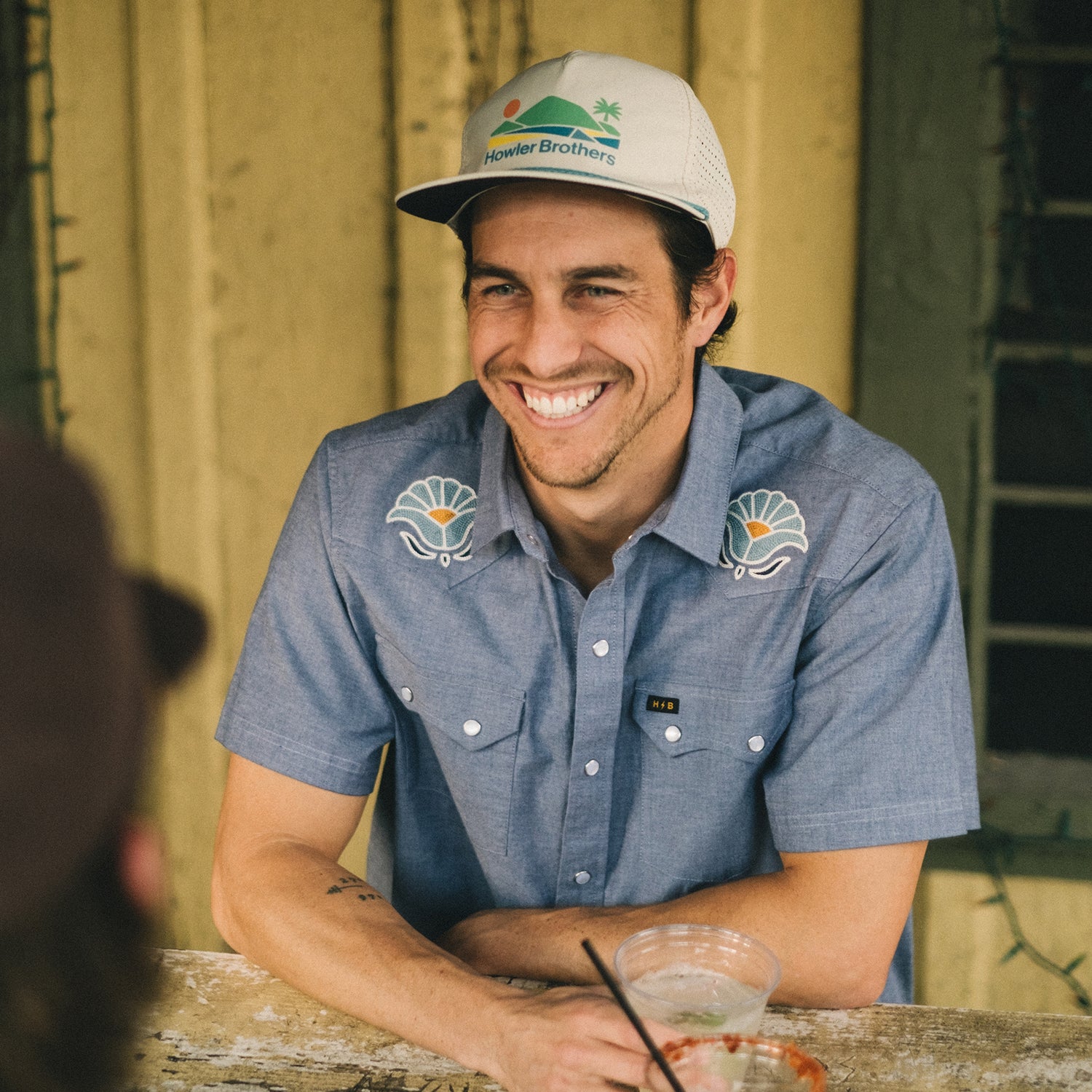 A man in a Crosscut Deluxe Shortsleeve Shirt - Fresh Blooms : Indigo and a trucker hat smiles while sitting at an outdoor wooden table with a drink in front of him.
