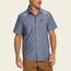 Man wearing a blue Crosscut Deluxe Shortsleeve summer shirt with motif embroidery of seashells on the shoulders, paired with brown pants, standing against a plain light background.