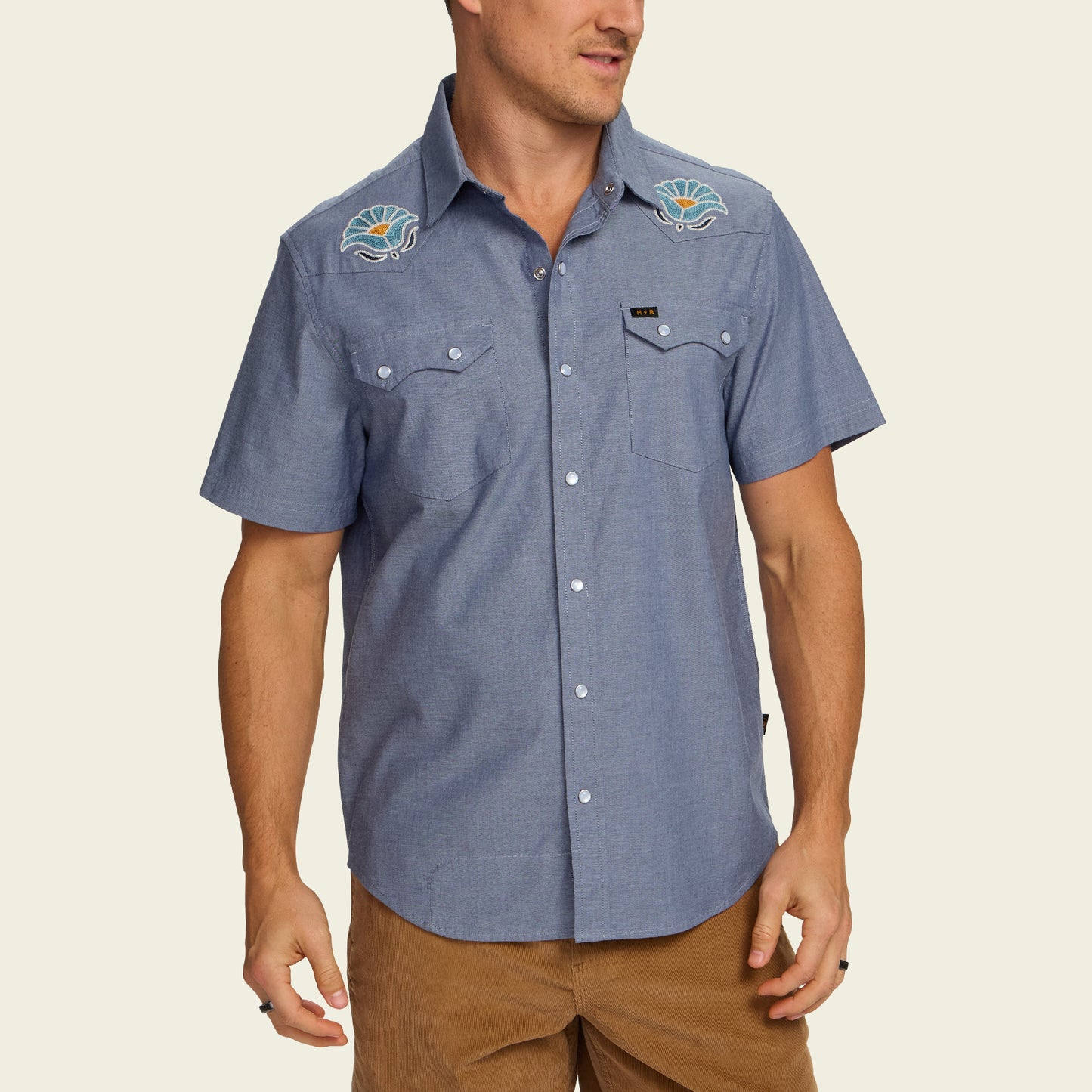 Man wearing a blue Crosscut Deluxe Shortsleeve summer shirt with motif embroidery of seashells on the shoulders, paired with brown pants, standing against a plain light background.