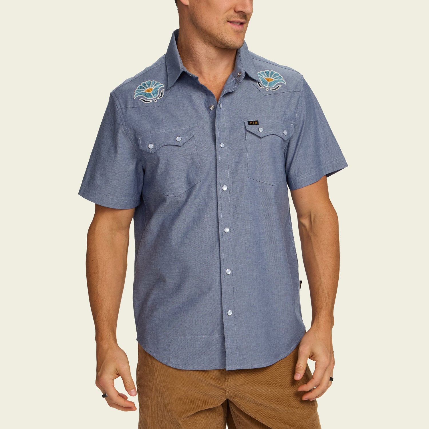 Man wearing a blue Crosscut Deluxe Shortsleeve summer shirt with motif embroidery of seashells on the shoulders, paired with brown pants, standing against a plain light background.