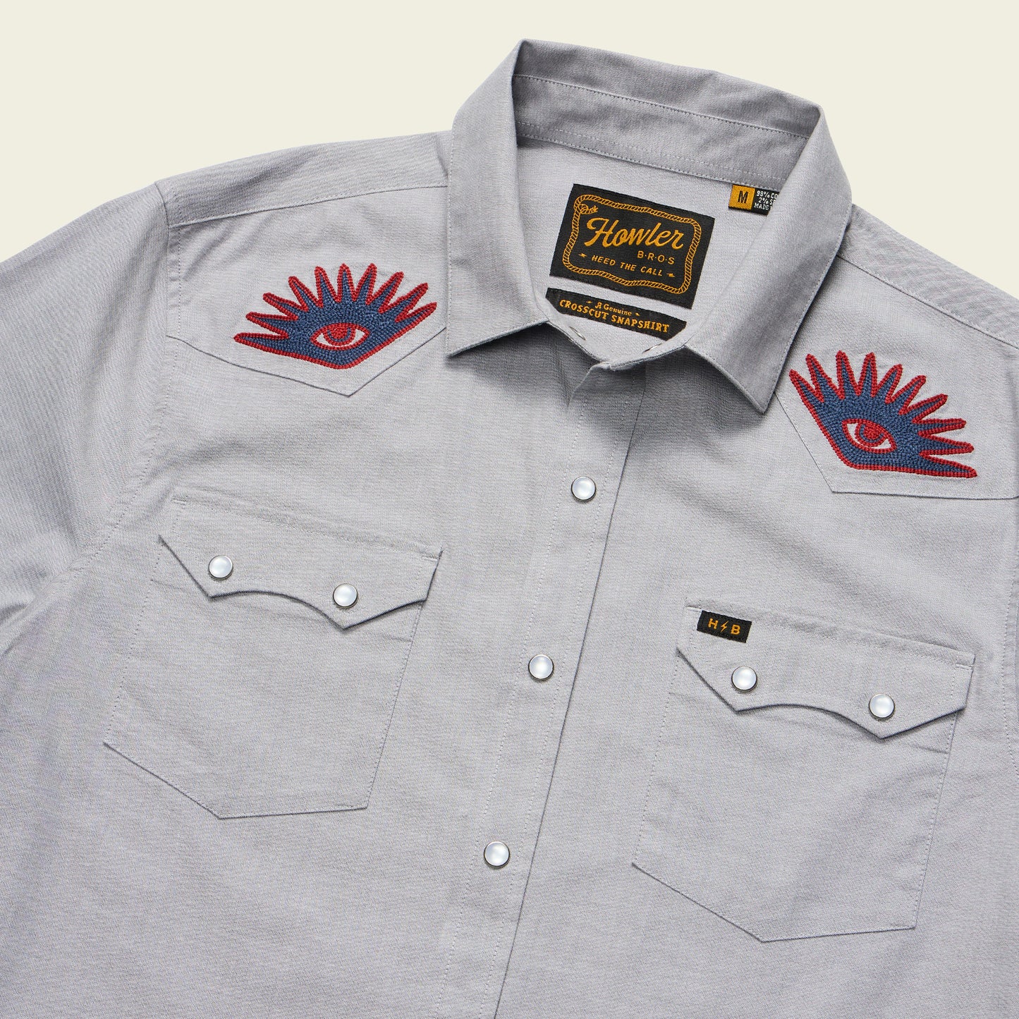A light gray Crosscut Deluxe Shortsleeve summer shirt with pearl snap buttons, two chest pockets, and red and blue motif embroidery of eye designs on both shoulders. The label reads Howler and indicates size Medium.