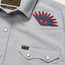 Close-up of a light gray Crosscut Deluxe Shortsleeve summer shirt with white snap buttons, a red and blue motif embroidery eye patch on the shoulder, and a small black H&B tag on the front pocket.