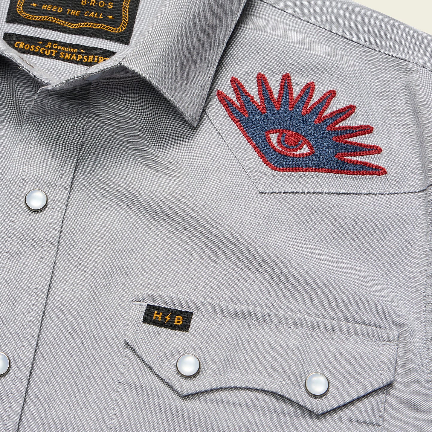 Close-up of a light gray Crosscut Deluxe Shortsleeve summer shirt with white snap buttons, a red and blue motif embroidery eye patch on the shoulder, and a small black H&B tag on the front pocket.