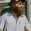 A bearded man wearing sunglasses, a trucker hat, and a Crosscut Deluxe Shortsleeve summer shirt with red motif embroidery on the shoulders stands outdoors in front of a weathered wooden wall.
