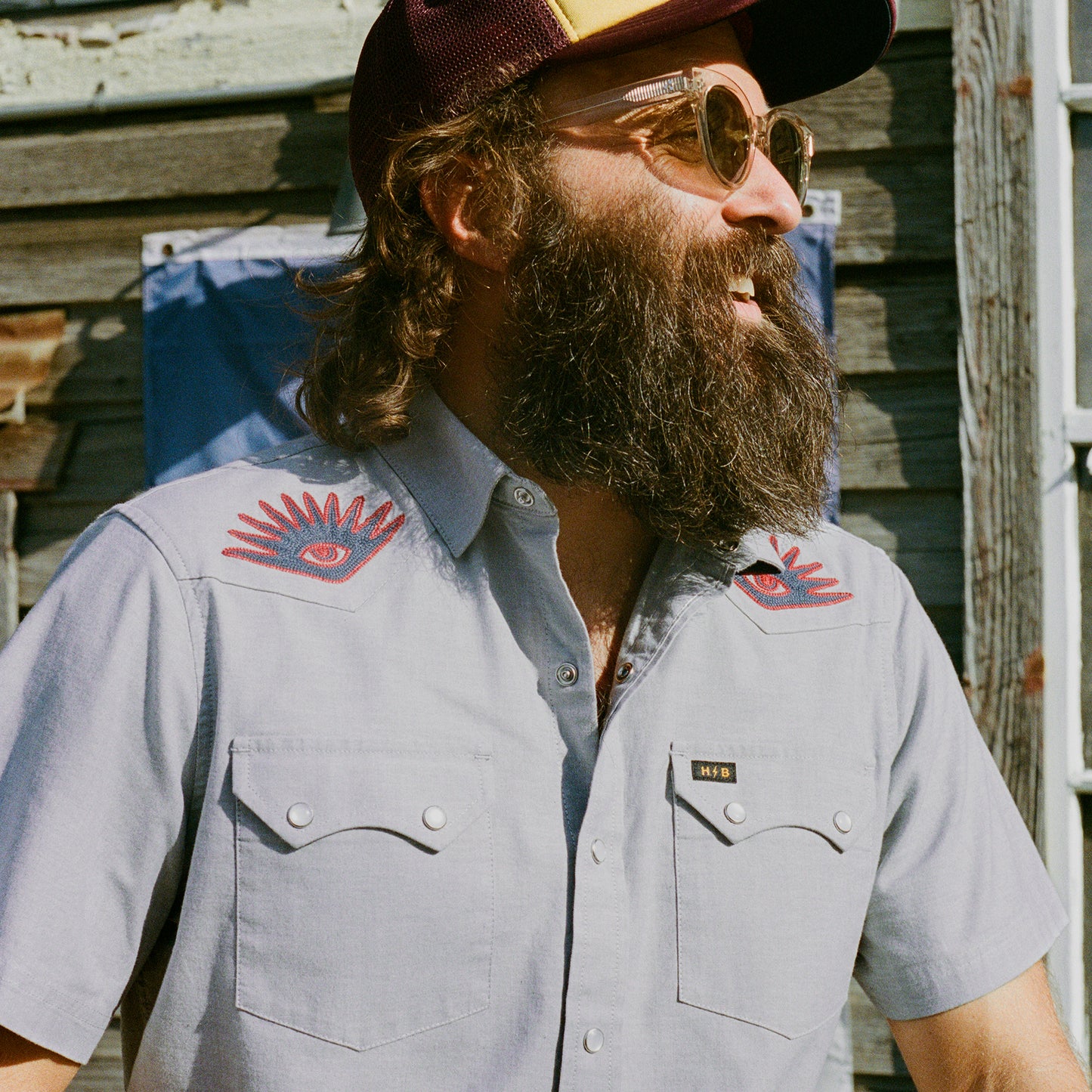 A bearded man wearing sunglasses, a trucker hat, and a Crosscut Deluxe Shortsleeve summer shirt with red motif embroidery on the shoulders stands outdoors in front of a weathered wooden wall.
