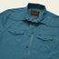 A teal lightweight button-up tech shirt with snap buttons, two chest pockets with flaps, and a visible Howler brand label sewn inside the collar area. Features UPF 35 protection and an HPL tag on one pocket. Shirt is laid flat on a light background.