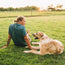A person in a lightweight tech shirt with blond hair sits on grass next to a large, light brown and white dog. They face each other on a sunny day in an open field with trees and a fence in the background.