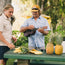 Two men stand at an outdoor table with sliced pineapples. One man, wearing an H Bar B snapshirt with pearl snaps, strums a ukulele while the other reaches for a pineapple slice amid lush greenery and a relaxed tropical setting.