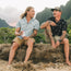 Two men with tattoos sit on a sandy ledge outdoors, smiling and talking. One sports an H Bar B Snapshirt with vintage inspired prints; both wear shorts, short-sleeved shirts, and enjoy scenic mountains and greenery in the background.