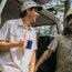 Two men in Howler Brothers vintage inspired shirts and caps laugh together by an open van door in a wooded area. One holds a blue mug, while the other shows a tattoo on his forearm as sunlight filters through the trees.