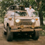 A person in a Howler Brothers pearl snap shirt and cap stands smiling next to an old, mud-covered Toyota SUV with surfboards on the roof, parked on a dirt road surrounded by green trees.