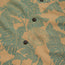 Close-up of a tan Monoloha Shirt with green tropical leaf patterns and brown buttons. The fabric features circular logos reading “Howler Brothers Overland Division Powered by Howler Land Cruiser.” Vintage Hawaiian shirts inspired.