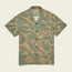 A short-sleeve, button-up Monoloha Shirt with a beige background and green leaf pattern, featuring a collar and front pocket. Inspired by vintage Hawaiian shirts, it offers a casual, tropical style perfect for any getaway.